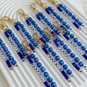 Op de afbeelding: Een verzameling sleutelhangers met blauwe gefacetteerde kralen, kleine goudkleurige tussenkralen en witte letterkralen die het woord MEMPHIS vormen. Elke sleutelhanger is bevestigd aan een goudkleurige karabijnsluiting voor een uniform geheel.