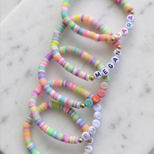 May include: Five stretch bracelets made of multicolored flat circular clay beads in pastel shades of pink, yellow, blue, green, and purple. Each bracelet features gold-toned spacer beads and white letter beads spelling the names Sara, Megan, Bella, Emily, and Ellie.