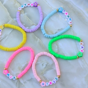May include: A collection of colorful beaded bracelets. Each bracelet features a different color, including yellow, pink, purple, blue, and green. The bracelets have white letter beads spelling out names and are accented with flower-shaped beads.