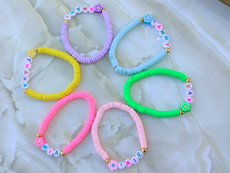 May include: A collection of colorful beaded bracelets. Each bracelet features a different color, including yellow, pink, purple, blue, and green. The bracelets have white letter beads spelling out names and are accented with flower-shaped beads.