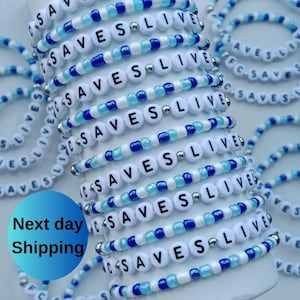May include: A collection of beaded bracelets. Each bracelet features white letter beads spelling "SAVES LIVE" interspersed with blue and light blue beads. The bracelets are stacked and arranged, showcasing the design and message.
