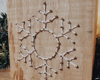 Peut inclure: Un flocon de neige en ficelle blanche sur une planche en bois. Le flocon de neige est réalisé avec de la ficelle blanche enroulée autour de petites punaises dorées.