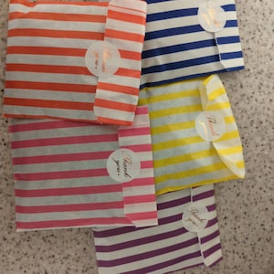 May include: A selection of striped paper gift bags in shades of orange, blue, yellow, pink, and purple. Each bag is sealed with a sticker that reads "Thank you". The bags are arranged on a light-colored surface.