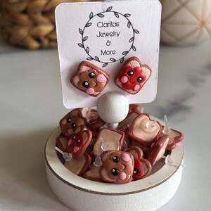 May include: A collection of handmade polymer clay earrings shaped like a slice of bread with a cute cartoon face. The earrings are displayed on a white stand with a sign that reads "Claritas Jewelry & More".