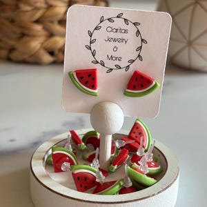 Könnte beinhalten: Ein weißer Präsentationsständer mit einem Schild mit der Aufschrift "Claritas Jewelry & More" und einer Auswahl an Wassermelonen-Schnitz-Ohrringen. Die Ohrringe sind rot und grün und haben eine weiße Rückseite.