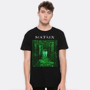 May include: Black t-shirt featuring the word "MATRIX" in white above a green graphic from the movie. The graphic shows a hallway with green lines and three figures. The t-shirt is made of a soft material.