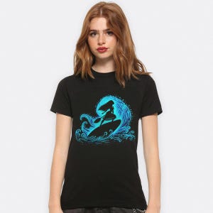 Puede incluir: Camiseta negra con un dise&ntilde;o azul de una persona surfeando una ola. La silueta del surfista se destaca contra una ola azul detallada. El dise&ntilde;o est&aacute; centrado en la camiseta.