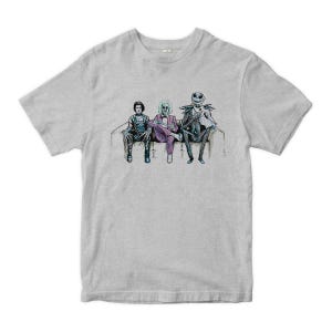 K&ouml;nnte beinhalten: Ein graues T-Shirt mit einem grafischen Aufdruck von drei Figuren, die auf einer Couch sitzen. Die Figuren sind in verschiedenen Kost&uuml;men gekleidet und scheinen aus einem Horrorfilm zu stammen.