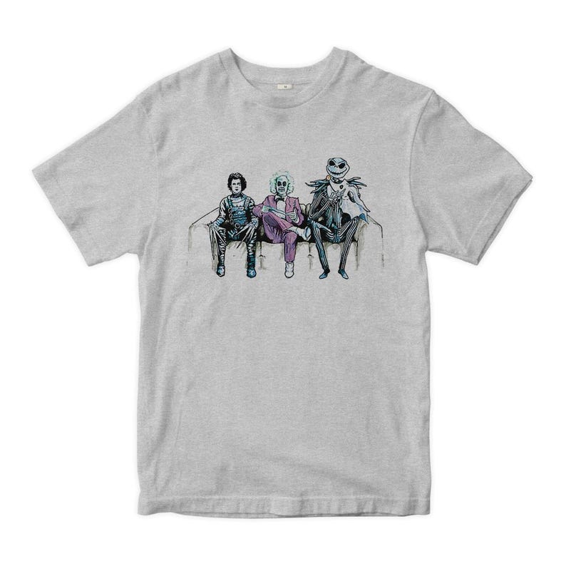 K&ouml;nnte beinhalten: Ein graues T-Shirt mit einem grafischen Aufdruck von drei Figuren, die auf einer Couch sitzen. Die Figuren sind in verschiedenen Kost&uuml;men gekleidet und scheinen aus einem Horrorfilm zu stammen.