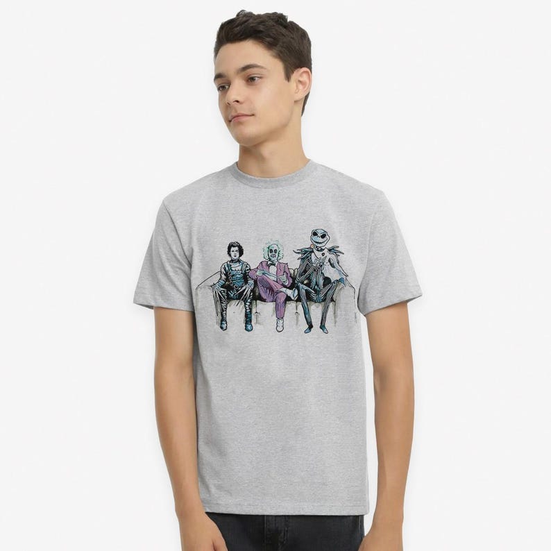 K&ouml;nnte beinhalten: Ein hellgraues T-Shirt mit einem Grafikdruck von drei Figuren aus dem Film Nightmare Before Christmas, die auf einer Couch sitzen. Die Figuren sind Jack Skellington, Sally und Oogie Boogie.