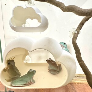 以下が含まれることがあります： 2つの白いセラミック製の雲の形をした囲いの中に、緑色の木登りガエルが4匹います。囲いは吸盤で天井から吊り下げられています。