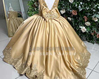 May include: A gold satin dress with a large bow and lace trim. The dress has a V-neckline and a full skirt. The dress is labeled "Lady Couture by Serap Ladim".