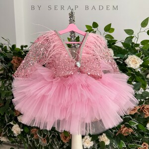 May include: A pink tulle tutu dress with a sequined bodice and feather trim. The dress is on a white mannequin and has a V-neckline. The text "Lady Couture by Serap Badem" is visible at the top of the image.