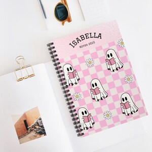 Könnte beinhalten: Ein rosa-weiß kariertes Notizbuch mit dem Text "ISABELLA NOTES 2025" auf dem Cover. Das Notizbuch ist mit einem Muster aus weißen Geistern, die Bücher lesen, und weißen Blumen verziert.