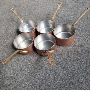 May include: A set of five round saucepans with copper-toned exteriors and silver-toned interiors. Each pan features a long, slender metal handle attached with two rivets. The pans are arranged in a circular pattern with handles pointing outwards.