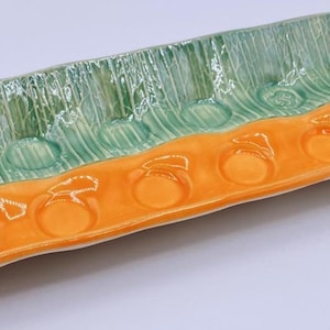 May include: A rectangular ceramic serving tray featuring a split color design with a glossy green textured section on one side and a smooth orange section on the other. The tray includes a row of six circular indentations along the orange side.