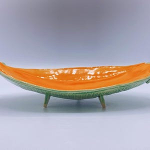 May include: An elongated ceramic bowl with a vibrant orange interior and a textured, light green exterior that mimics the appearance of a leaf. The vessel is supported by three small, short legs and features a curved, boat-like shape.