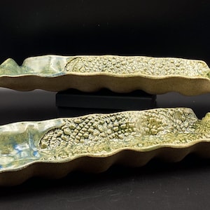 Pode incluir: Duas travessas de cerâmica feitas à mão. As travessas são longas e onduladas, com uma superfície texturizada e salpicada em tons de verde e bege. As bordas têm um design ondulado. As travessas são exibidas em uma superfície preta.