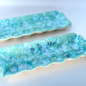 Peut inclure: Deux plateaux rectangulaires en céramique aux bords ondulés, dotés d'un émail brillant turquoise et blanc tacheté. Le plateau avant présente un petit motif bleu foncé représentant un oiseau et du feuillage, le second a un motif uniforme.