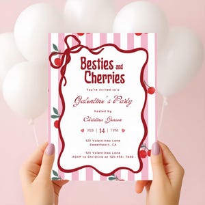 以下が含まれることがあります： 「Besties and Cherries」と「Galentine's Party」の文字が入った、ピンクと白のストライプの招待状。 招待状は両手で持たれており、チェリーのデザインと2月14日の日付が特徴です。