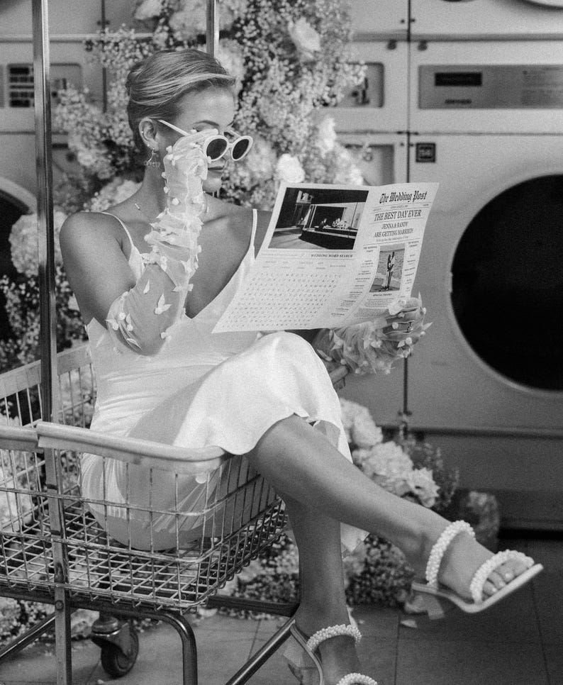 May include: A black and white image of a woman in a white dress and sunglasses reading a newspaper titled "The Wedding Post". She is sitting in a shopping cart, with floral arrangements in the background. She is wearing white gloves and pearl-adorned sandals.
