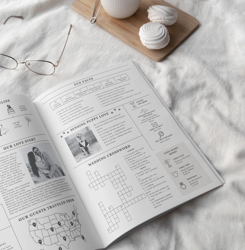 May include: An open wedding journal with text and photos, featuring a crossword puzzle, fun facts, and a map of guest locations. A wooden cutting board with a white mug and two meringues is in the background.