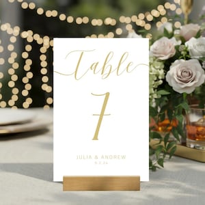 May include: A white rectangular table number card featuring the gold text Table 7, with the names Julia & Andrew and the date 9.2.24 printed below in a smaller font, held upright by a rectangular wooden base stand on a light-colored surface.