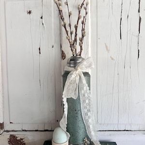 Puede incluir: Arreglo de estilo vintage con un jarr&oacute;n de metal verde azulado, borde plateado, cinta de encaje crema y ramas de sauce. Un huevo azul claro descansa en un peque&ntilde;o recipiente blanco, sobre un libro verde. El jarr&oacute;n mide unos 20 cm de alto.