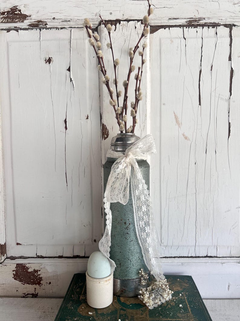 Puede incluir: Arreglo de estilo vintage con un jarr&oacute;n de metal verde azulado, borde plateado, cinta de encaje crema y ramas de sauce. Un huevo azul claro descansa en un peque&ntilde;o recipiente blanco, sobre un libro verde. El jarr&oacute;n mide unos 20 cm de alto.