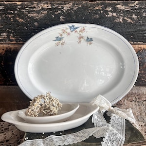 May include: A collection of vintage white ceramic serving dishes. The large oval platter features a blue rim and a floral and bird design. Two smaller dishes and a decorative lace ribbon are also present.
