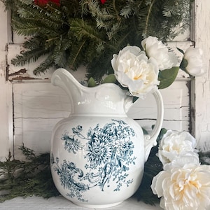 Może przedstawiać: Biały ceramiczny dzban z niebiesko-zielonym kwiatowym wzorem. Dzban wypełniony jest białymi piwoniami. Zielony wieniec i czerwona kokarda w tle. Przedmiot dekoracyjny.