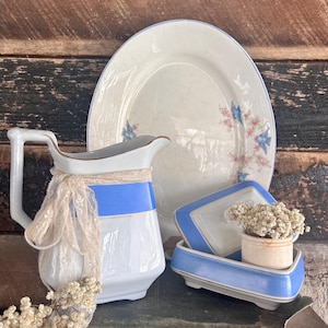 Puede incluir: Una colección de artículos de cerámica vintage blancos y azules, que incluyen una jarra con encaje, una fuente para servir y un jabonera. El plato tiene un diseño floral. Los artículos están dispuestos sobre una pila de libros antiguos.