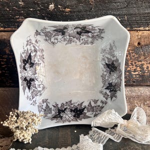 May include: A white, square dish with a decorative black and grey leaf design around the rim. The dish is displayed on a dark wooden surface, with dried flowers and lace trim.