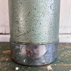 Puede incluir: Un recipiente met&aacute;lico cil&iacute;ndrico vintage con un acabado texturizado y moteado de color verde claro. La base es de metal brillante de color plateado. El recipiente descansa sobre una superficie verde estampada, con peque&ntilde;as flores blancas esparcidas.