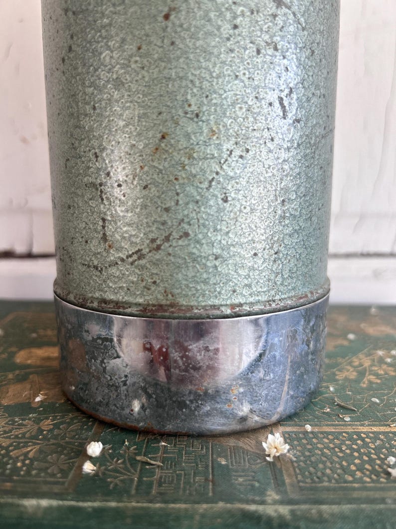 Puede incluir: Un recipiente met&aacute;lico cil&iacute;ndrico vintage con un acabado texturizado y moteado de color verde claro. La base es de metal brillante de color plateado. El recipiente descansa sobre una superficie verde estampada, con peque&ntilde;as flores blancas esparcidas.