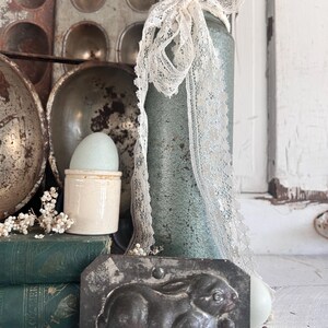 Puede incluir: Naturaleza muerta r&uacute;stica con elementos de Pascua. Molde de conejo de metal, tarro azul-verdoso con encaje y huevera de cer&aacute;mica con huevo, dispuestos con libros antiguos y moldes para hornear. La escena evoca una est&eacute;tica de granja.