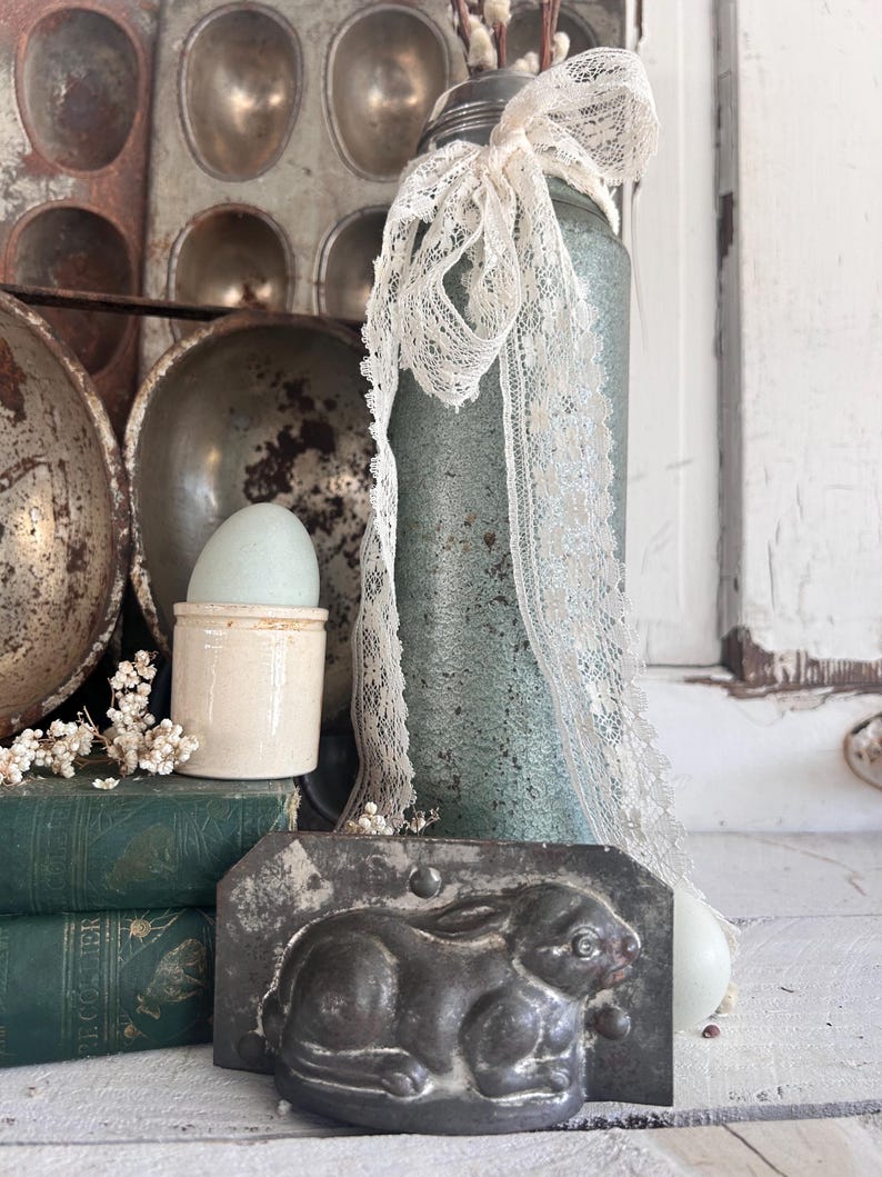 Puede incluir: Naturaleza muerta r&uacute;stica con elementos de Pascua. Molde de conejo de metal, tarro azul-verdoso con encaje y huevera de cer&aacute;mica con huevo, dispuestos con libros antiguos y moldes para hornear. La escena evoca una est&eacute;tica de granja.