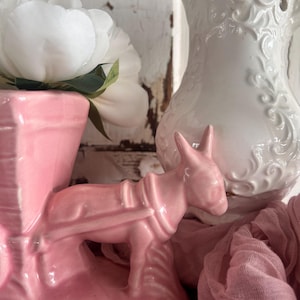 Könnte beinhalten: Eine rosa Keramik-Eselfigur mit Geschirr, vor dem Hintergrund einer weißen Pfingstrose und einem weißen, verzierten Krug. Der Esel ist der Mittelpunkt, mit detailliertem Design. Ein rosa Stoff fügt der Szene eine weiche Textur hinzu.