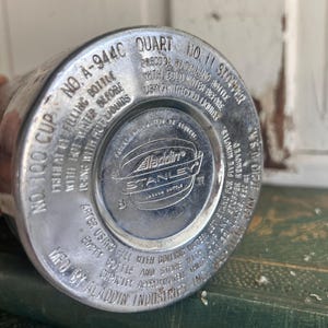 Puede incluir: Primer plano de la base de una botella de vac&iacute;o Aladdin Stanley vintage. La base de metal plateado presenta texto en relieve, incluyendo "Aladdin Industries Inc." y "Stanley Vacuum Bottle". La botella est&aacute; marcada como "1 Quart" y "No. 11 Stopper".
