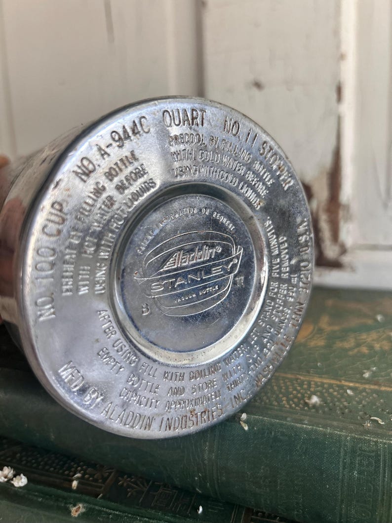 Puede incluir: Primer plano de la base de una botella de vac&iacute;o Aladdin Stanley vintage. La base de metal plateado presenta texto en relieve, incluyendo "Aladdin Industries Inc." y "Stanley Vacuum Bottle". La botella est&aacute; marcada como "1 Quart" y "No. 11 Stopper".