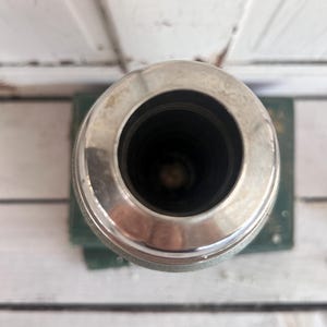 Puede incluir: Un recipiente cil&iacute;ndrico de color plateado con un borde ancho y brillante. El interior es oscuro y el recipiente se asienta sobre una base verde. La imagen est&aacute; tomada desde una perspectiva superior, destacando la abertura circular.