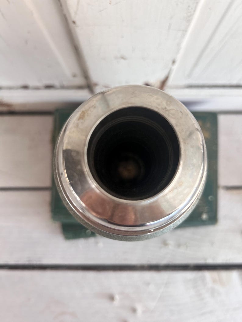 Puede incluir: Un recipiente cil&iacute;ndrico de color plateado con un borde ancho y brillante. El interior es oscuro y el recipiente se asienta sobre una base verde. La imagen est&aacute; tomada desde una perspectiva superior, destacando la abertura circular.
