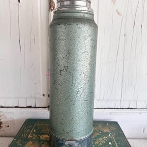 Puede incluir: Termo verde y plateado vintage. La botella cil&iacute;ndrica tiene un acabado texturizado y moteado. La parte superior e inferior son de metal plateado. El termo est&aacute; sobre un libro verde con letras doradas.