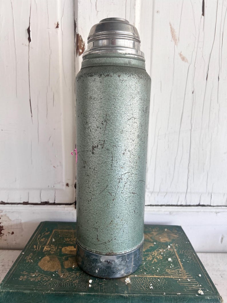 Puede incluir: Termo verde y plateado vintage. La botella cil&iacute;ndrica tiene un acabado texturizado y moteado. La parte superior e inferior son de metal plateado. El termo est&aacute; sobre un libro verde con letras doradas.