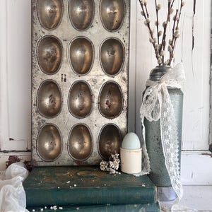 Puede incluir: Un molde antiguo de metal para hornear huevos con 12 cavidades ovaladas. El molde tiene una p&aacute;tina r&uacute;stica. Un jarr&oacute;n vintage con ramas de sauce y una cinta de encaje se encuentra junto al molde. Tambi&eacute;n hay dos libros verdes antiguos y un huevero.