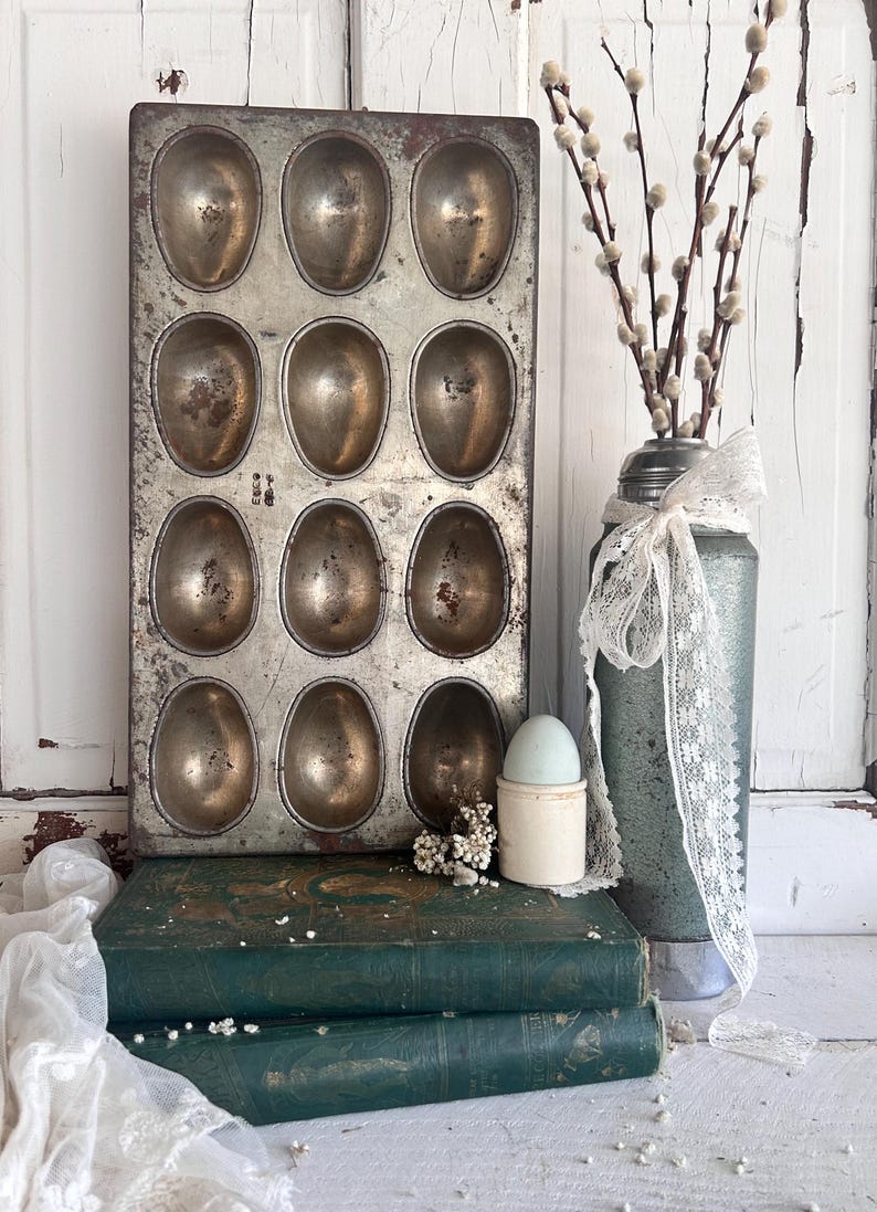 Puede incluir: Un molde antiguo de metal para hornear huevos con 12 cavidades ovaladas. El molde tiene una p&aacute;tina r&uacute;stica. Un jarr&oacute;n vintage con ramas de sauce y una cinta de encaje se encuentra junto al molde. Tambi&eacute;n hay dos libros verdes antiguos y un huevero.