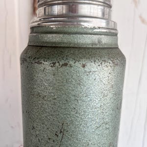 Puede incluir: Una coctelera de estilo vintage con un cuerpo verde p&aacute;lido texturizado y moteado y una tapa plateada brillante. La coctelera muestra signos de desgaste, lo que aumenta su encanto vintage. El dise&ntilde;o recuerda a la cristaler&iacute;a de bar cl&aacute;sica.