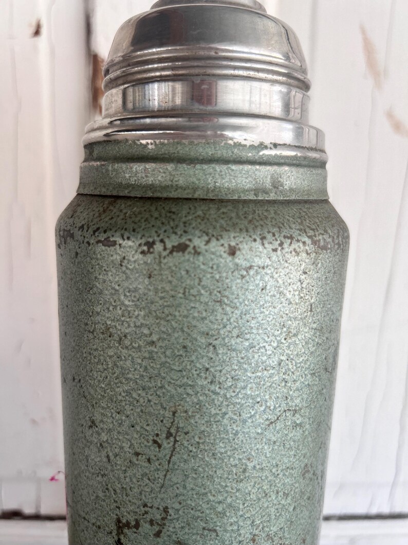 Puede incluir: Una coctelera de estilo vintage con un cuerpo verde p&aacute;lido texturizado y moteado y una tapa plateada brillante. La coctelera muestra signos de desgaste, lo que aumenta su encanto vintage. El dise&ntilde;o recuerda a la cristaler&iacute;a de bar cl&aacute;sica.