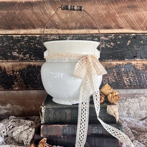 May include: A white ceramic vase with a metal handle and a cream-colored lace bow. The vase is decorated with a lace trim and sits atop a stack of antique books. Dried roses and lace fabric add to the vintage aesthetic.