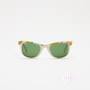 May include: Off-white sunglasses with green lenses and colorful patterned frames. The frames feature a mix of pink, green, and yellow hues. The sunglasses have a classic, rectangular shape and are set against a white background.